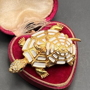 May include: A gold and white turtle brooch with rhinestones. The turtle is on top of another turtle. The brooch is in a red velvet heart-shaped box.