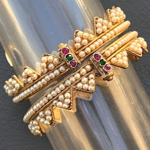 May include: Two gold-toned bangles with intricate designs. Each bangle features a row of small, white pearls and is accented with red and green gemstones. The bangles have a detailed, textured appearance, suggesting a vintage or antique style.