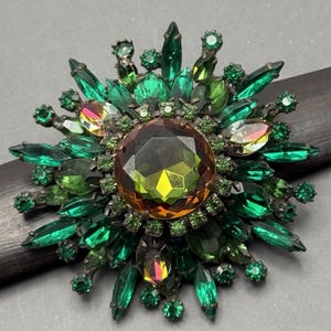 May include: A large, green and gold rhinestone brooch in the shape of a flower. The brooch has a large, faceted, amber-colored center stone surrounded by smaller, green rhinestones.