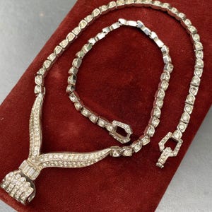May include: A silver rhinestone necklace with a V-shaped pendant. The necklace is on a red velvet background.