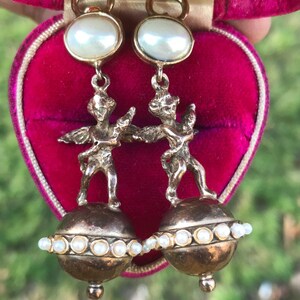 May include: A pair of gold tone earrings with a pearl drop and a cherub figure standing on a sphere of pearls.