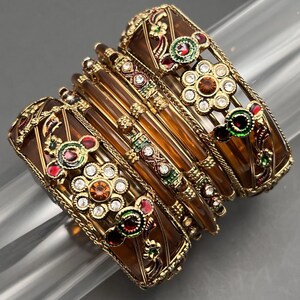 Könnte beinhalten: Ein Set aus sechs goldfarbenen Armreifen mit braunen Glasböden. Die Armreifen sind mit buntem Emaille und klaren und bernsteinfarbenen Strasssteinen verziert.