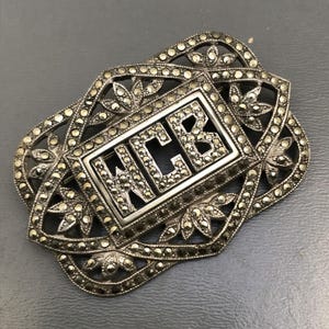 May include: A silver brooch with a rectangular frame set with marcasite stones. The frame contains the letters "HCB" also set with marcasite stones.