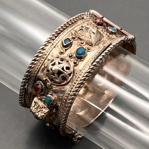 Può includere: Bracciale a polsino in argento con dettagli intricati, ornato da pietre preziose rosse e turchesi. Il bracciale presenta un bordo intrecciato e un'elaborata lavorazione del metallo, che mostra un design vintage o etnico. Il bracciale è esposto su un oggetto cilindrico trasparente.