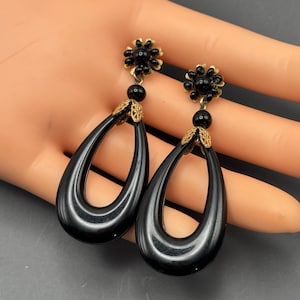 Puede incluir: Un par de pendientes negros de gota con detalles dorados. Los pendientes tienen una forma ovalada negra con un diseño de filigrana dorada y una parte superior en forma de flor negra.