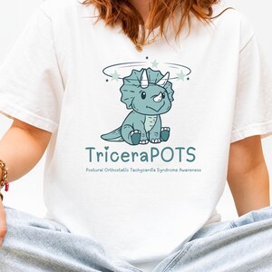 Pode incluir: Camiseta branca com um desenho de Triceratops de desenho animado. O texto diz "TriceraPOTS" e "Conscientização sobre a síndrome da taquicardia ortostática postural" em verde-azulado. O dinossauro é azul claro com um halo e estrelas.