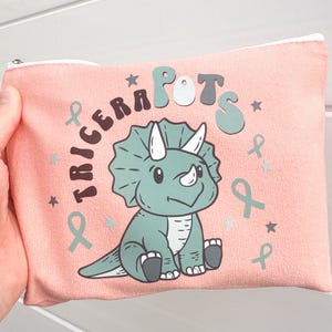 May include: A pink canvas zippered pouch with a green triceratops illustration and the text "Triceratops" and "Pots" in a colourful, stylised font.