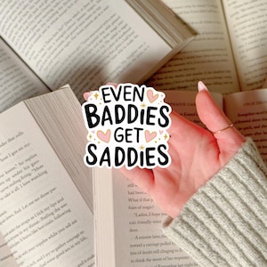 May include: A sticker with the text "EVEN BADDIES GET SADDIES" in black, with pink hearts and gold stars. The sticker is held over an open book with visible text. The background includes other open books.