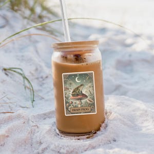 May include: A glass tumbler with a wooden lid and a clear straw, filled with a light brown beverage. A sticker on the glass features a green frog on a mushroom, with the text "BRAIN FROG" and a crescent moon.