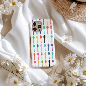 May include: White iPhone case with a colorful spoon pattern. The design features rows of spoons in various colors, including red, yellow, green, and purple. The case is surrounded by white fabric and daisies.