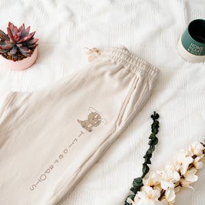 May include: Cream-colored sweatpants with a cartoon dinosaur graphic and the text "Tri-ce-tops Sports". A green and white mug with the words "Home Heart & Soul" is next to the pants. A succulent in a pink pot and flowers are also in the image.