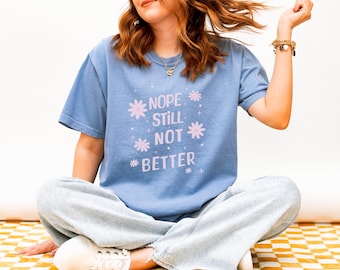 Op de afbeelding: Lichtblauw T-shirt met de tekst "Nope Still Not Better" in wit en roze, samen met bloem- en sterrenafbeeldingen. Het shirt wordt gedragen met lichtblauwe jeans.