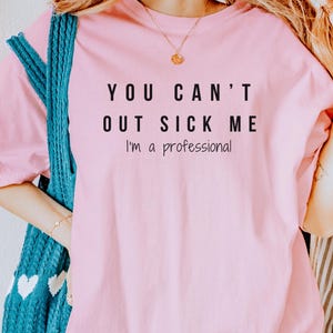 Puede incluir: Camiseta rosa con el texto "YOU CAN'T OUT SICK ME I'm a professional" impreso en negro.