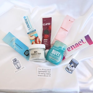 May include: An assortment of hydration products, including electrolyte drink mixes, hydration drops, and sea salt, are arranged on a white satin surface. The products feature various flavors and brands, with packaging in shades of blue, pink, and white. Stickers with text are also included.