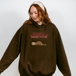 Op de afbeelding: Een donkerbruine hoodie met een kangoeroezak. De voorkant van de hoodie heeft de tekst "PERSONALLY VICTIMIZED BY MY IMMUNE SYSTEM" in roze, met een cartoon illustratie van een luiaard eronder.