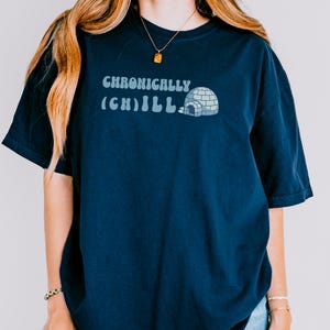 Peut inclure: T-shirt bleu marine avec l'inscription "Chronically (Ch)ill" et un dessin d'igloo bleu clair. Le t-shirt est de coupe ample, adapté aux tenues décontractées.