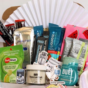 May include: A gift basket filled with various hydration and electrolyte products. Includes packets of Pedialyte, Liquid I.V., and Salt Stick chews, along with a cooling patch, stickers, and a pair of watermelon-themed socks. The basket is arranged in front of a white paper fan.