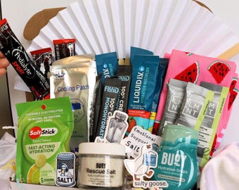 May include: A gift basket filled with various hydration and electrolyte products. Includes packets of Pedialyte, Liquid I.V., and Salt Stick chews, along with a cooling patch, stickers, and a pair of watermelon-themed socks. The basket is arranged in front of a white paper fan.