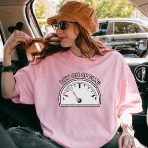 May include: A pale pink t-shirt featuring the words "LOW ON SPOONS" above a gauge graphic. The gauge shows a spoon and needle pointing towards the empty side. The person in the image is wearing sunglasses and a brown hat.