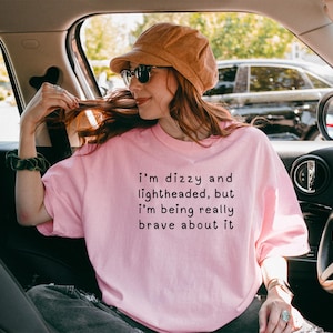 May include: A pink t-shirt with the text "I'm dizzy and lightheaded, but I'm being really brave about it" printed on the front. The person wearing the shirt is sitting in a car.