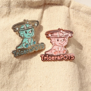 May include: Two enamel pins featuring a cartoon Triceratops with a star-shaped halo. The pins are in a silver and gold finish. The text "TriceraPOTS" is on the bottom of each pin.