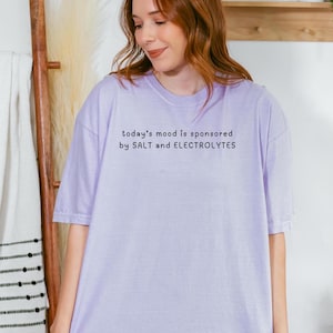 May include: A lavender t-shirt with the text "today's mood is sponsored by SALT and ELECTROLYTES" printed in black.