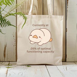 May include: A white tote bag with a cartoon illustration of a yellow chick lying on its back. The text on the bag reads "Currently at .05% of optimal functioning capacity."