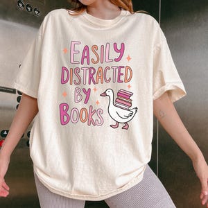 Peut inclure: Un t-shirt blanc avec le texte "Easily Distracted by Books" en lettres roses et orange. Un dessin animé d'une oie portant une pile de livres se trouve sous le texte.