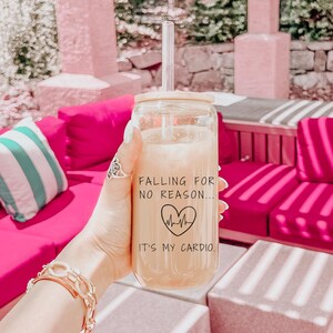 May include: A clear glass tumbler with a bamboo lid and a straw, filled with a light-colored beverage. The tumbler has the text "Falling for no reason... It's my cardio" with a heart and heartbeat graphic. The tumbler is held in a hand.