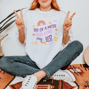 May include: White t-shirt with the text "Bit of a mess but doin' my best" in purple, orange, and yellow. The shirt features a rainbow graphic. The person is wearing blue jeans and white trainers.
