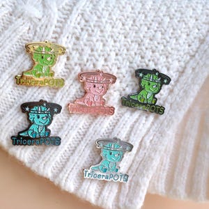 Könnte beinhalten: Fünf Emaille-Pins mit einem Cartoon-Triceratops, der eine Krone mit Sternen trägt. Jeder Pin hat ein anderes Farbschema: grün, rosa, blau, türkis und schwarz. Die Pins tragen unten den Schriftzug "TriceraPOTS".