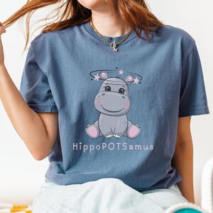 Puede incluir: Una camiseta azul con un hipopótamo de dibujos animados que lleva un halo con estrellas. El texto "Hippopotamus" está impreso debajo del hipopótamo.