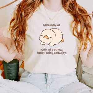 Könnte beinhalten: Ein weißes T-Shirt mit einer Cartoon-Illustration eines gelben Küken mit dem Text "Aktuell bei 0,05 % der optimalen Funktionsfähigkeit".