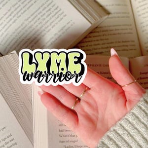 Peut inclure: Un autocollant avec les mots "LYME warrior" dans une police rétro, soulignés en noir et remplis d'une couleur vert clair. L'autocollant est tenu devant des livres ouverts.