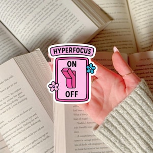 Op de afbeelding: Een roze sticker met de woorden "HYPERFOCUS", "ON" en "OFF" in een rechthoekig ontwerp. De sticker heeft een tuimelschakelaar-afbeelding en kleine bloemenillustraties. De sticker wordt boven een open boek gehouden.