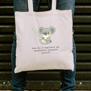 May include: A white tote bag with a cartoon koala holding a spoon. The text on the bag reads "How do I register my emotional support spoon?"