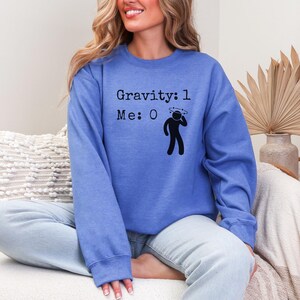 以下が含まれることがあります： 「Gravity: 1 Me: 0」という文字と、めまいがする顔をした棒人間が描かれた青いスウェットシャツです。