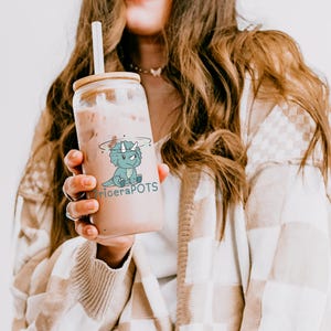 May include: A clear glass reusable bottle with a bamboo lid and a straw. The bottle has a cartoon dinosaur graphic with the text "TriceraPOTS".