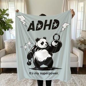 May include: A light blue throw blanket featuring a black and white panda illustration lifting a kettlebell. The text "ADHD" is written in black with white stars on either side. The text "It's my superpower." is written in black at the bottom of the blanket.
