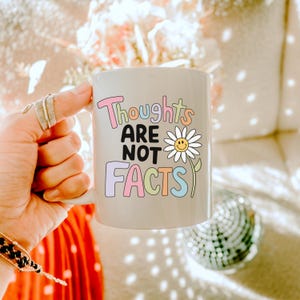 May include: A white ceramic mug with the text "Thoughts are not facts" in colorful letters and a daisy with a smiley face.