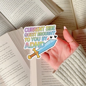 Könnte beinhalten: Ein farbenfroher Aufkleber mit dem Text "CURRENT SIDE QUEST BROUGHT TO YOU BY ADHD" und einem Cartoon-Schwert. Der Aufkleber hat einen Regenbogen-Farbverlauf-Hintergrund und wird über einem offenen Buch gehalten.