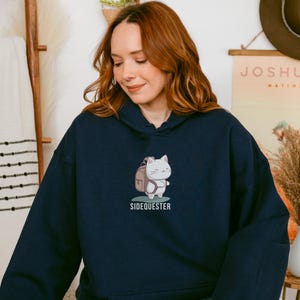 May include: A navy blue hoodie with a white cat wearing a backpack and the text "Sidequester" printed on the front.