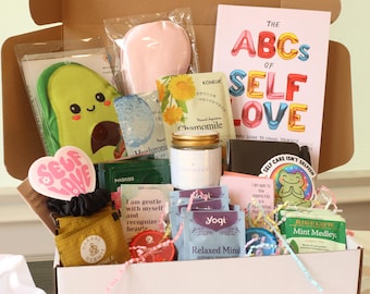 May include: A gift box filled with self-care items. Includes a book titled "The ABCs of Self Love", a plush avocado, tea bags, a jar of lavender, face masks, and a heart-shaped sticker that says "Self Love".