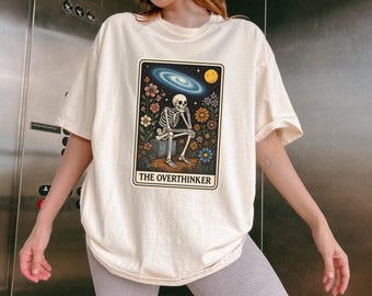 Puede incluir: Camiseta color crema con un diseño de carta del tarot que presenta un esqueleto, flores y elementos celestiales. La carta dice "THE OVERTHINKER". La camiseta tiene mangas cortas y un corte holgado.