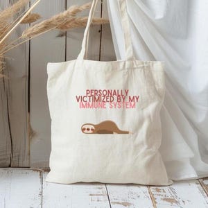 May include: A white canvas tote bag with the text "PERSONALLY VICTIMIZED BY MY IMMUNE SYSTEM" in pink and red. A cartoon sloth is printed below the text.