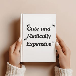 May include: A white book with the text "Cute and Medically Expensive" in black, along with small pill illustrations. The book is held by two hands with manicured nails, against a neutral background.