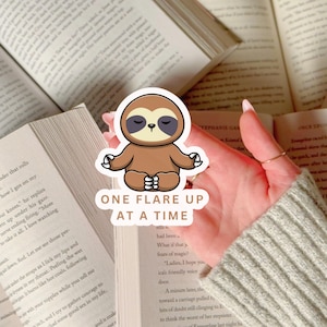 May include: A sticker featuring a cartoon sloth in a meditative pose, colored in shades of brown and beige. The sticker has the text "ONE FLARE UP AT A TIME" in a white font. The sticker is placed on top of open books.