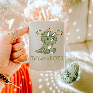 Puede incluir: Taza de cerámica blanca con un triceratops de dibujos animados verde que lleva una corona de estrellas. La taza tiene el texto "TriceraPOTS" impreso en la parte delantera.