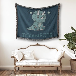 May include: A blue and green tapestry with a cartoon triceratops wearing a halo of stars. The tapestry has a fringe border and the text "TriceraPOTS" is printed on the bottom.