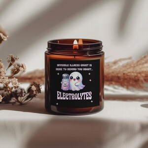 May include: A brown glass candle jar with a lit wick. The label features a cartoon ghost and salt shaker with the text "INVISIBLE ILLNESS GHOST IS HERE TO REMIND YOU ABOUT... ELECTROLYTES."